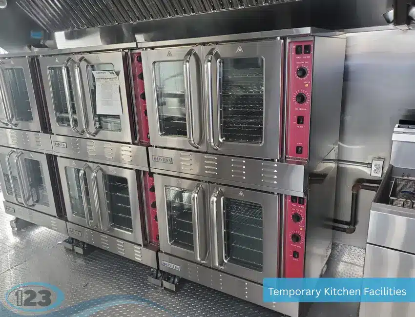 A row of professional double-stacked Kintera stainless steel convection ovens with red control panels inside a temporary kitchen facility featuring an overhead hood and diamond plate flooring.