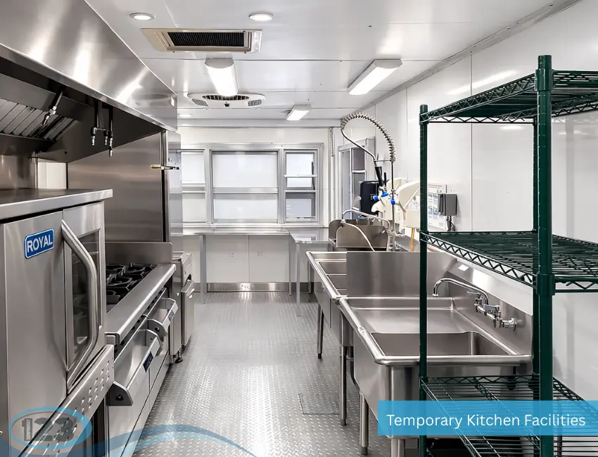 Interior view of a portable commercial kitchen featuring stainless steel cooking equipment, double sinks, industrial stove, shelving, and clean stainless steel work surfaces in a compact trailer-style layout.