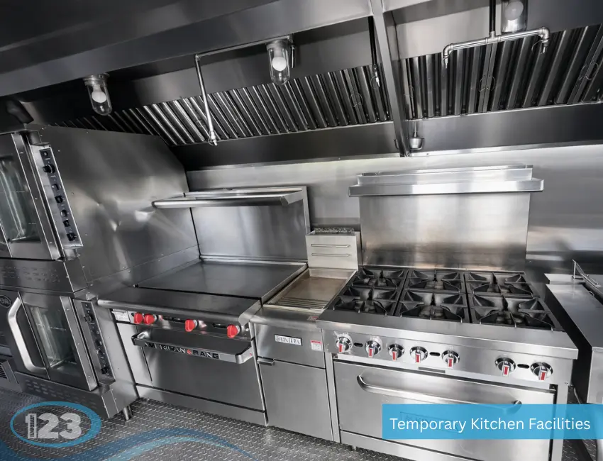 A commercial hot line inside a temporary kitchen facility featuring an American Range flat-top griddle with red knobs, a Kintera 6-burner gas stove, double-stacked ovens, and a massive stainless steel exhaust hood overhead.
