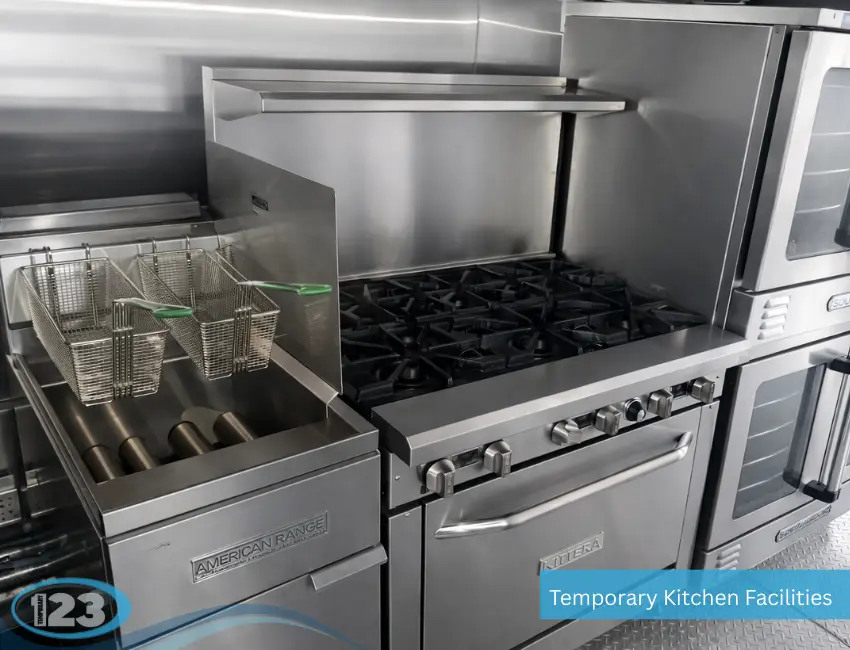A stainless steel commercial cooking line inside a temporary kitchen facility featuring an American Range deep fryer with green-handled wire baskets, a Kintera six-burner gas stovetop, and double-stacked Southbend ovens on the side.
