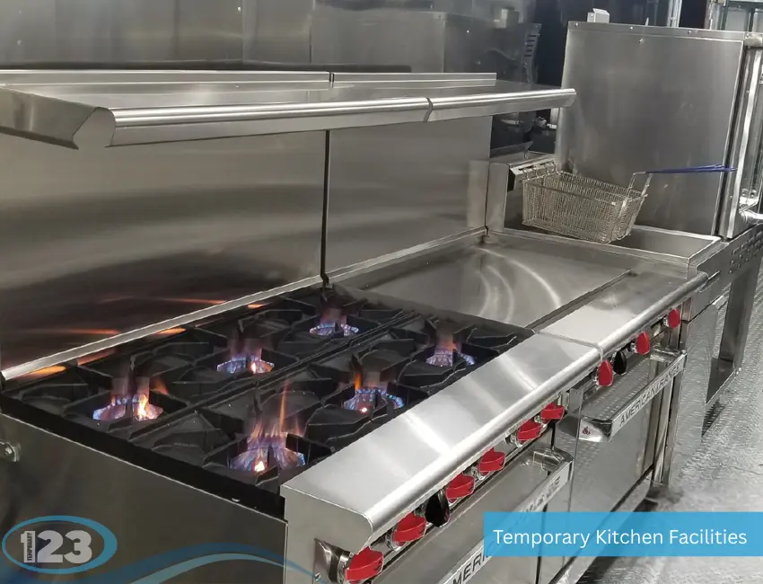 Close-up of an active commercial hot line inside a temporary kitchen facility, showcasing an American Range six-burner gas stovetop with lit blue flames, a flat-top griddle, and an industrial deep fryer in the background.