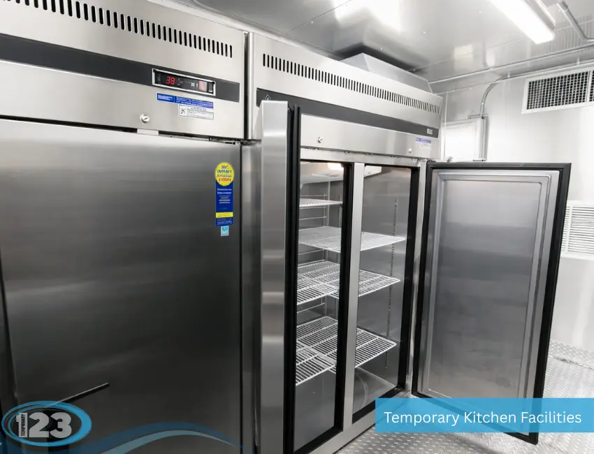 Interior view of a temporary kitchen facility featuring two large stainless steel commercial reach-in refrigerators, with one solid door unit on the left showing a digital temperature display of 38 degrees, and one open glass-door unit on the right showing empty wire racks.