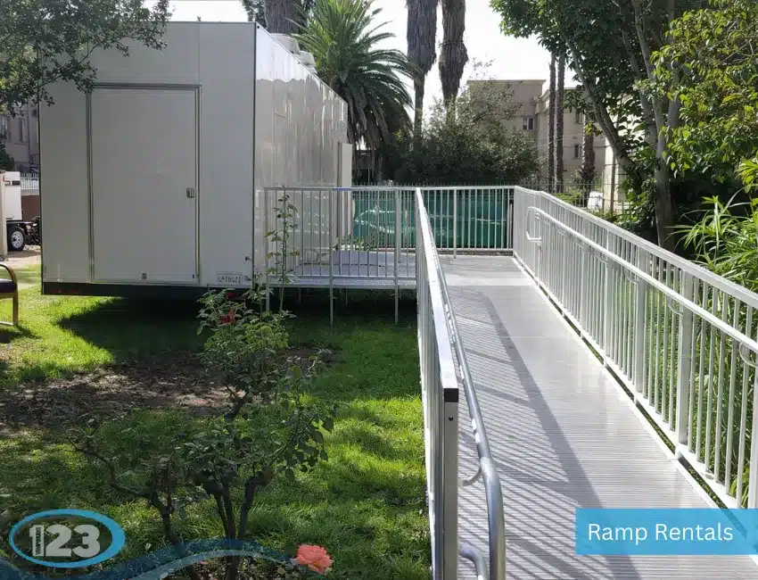 An outdoor shot of a long, temporary aluminum modular ramp with handrails leading up to the entrance platform of a white mobile trailer unit parked on a grassy lawn.