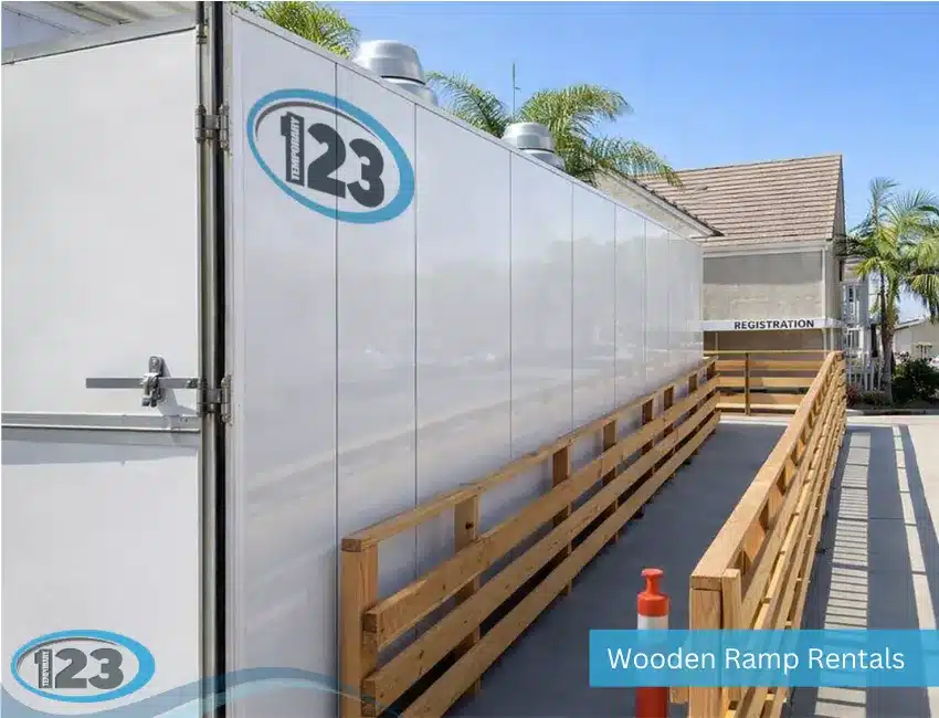 An outdoor wooden accessibility ramp with double handrails installed alongside a large white mobile modular unit with a "123 Temporary" logo under a clear blue sky.